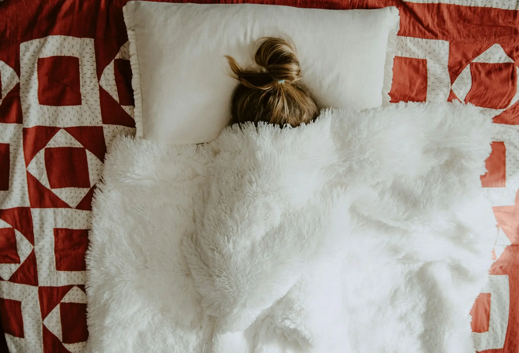 Person resting under a fluffy white throw on a patchwork quilt